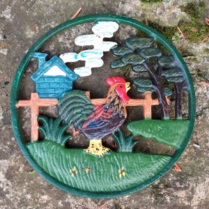 Peut inclure: Un dessous de plat en fonte verte avec un coq, une maison et un arbre. Le coq est rouge, jaune et brun. La maison est verte et blanche. L'arbre est vert.