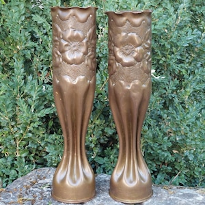 May include: Two tall, cylindrical copper-toned metal vases featuring a scalloped top edge and a relief design of embossed flowers and leaves. The lower sections of the vases have vertical fluted grooves that taper toward the base.