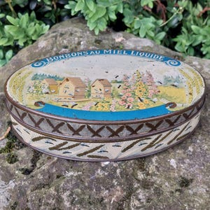 Peut inclure: Boîte ovale vintage avec couvercle orné d'une scène champêtre avec des ruches, des fleurs et des abeilles. La boîte a un bord bleu et le texte "BONBONS AU MIEL LIQUIDE" sur le dessus. Les côtés présentent un motif répété de blé et d'insectes.