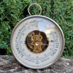 Può includere: Un barometro aneroide rotondo color argento con quadrante bianco, testo nero e un meccanismo centrale in ottone. Il quadrante presenta indicazioni meteorologiche in francese tra cui Tempête, Pluie, Variable, Beau Temps e Très Sec, con anello.