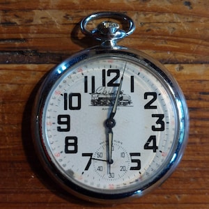 Puede incluir: Un reloj de bolsillo plateado con esfera blanca y números negros. La esfera del reloj presenta la palabra "Chile" sobre una ilustración de un tren. El reloj tiene un anillo en la parte superior para sujetar una cadena.