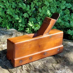 Peut inclure: Un rabot à main en bois avec une lame en fer brun foncé. Le rabot est en bois brun clair et comporte quatre vis sur le dessous.