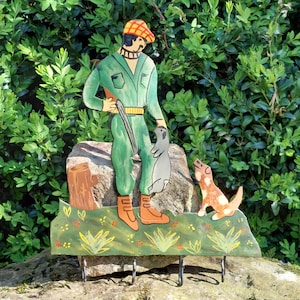 Peut inclure: Un porte-clés en bois peint représentant un chasseur en salopette verte, tenant un fusil et un animal capturé. Un chien est à côté. Le porte-clés a quatre crochets en métal et est placé sur un fond de feuilles vertes.