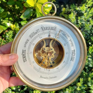 May include: A classic brass barometer with a white dial and black lettering. The dial displays readings such as "Variable", "Beau Fixe", and "Tempete". It features a glass cover and a hanging ring. The internal mechanism is visible.