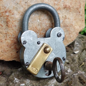 Peut inclure: Un cadenas vintage de couleur argent avec un loquet en laiton et une clé. Le cadenas a une forme arrondie unique et les lettres "V.D." sont gravées sur le loquet en laiton.