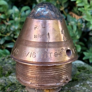 Puede incluir: Un proyectil de artillería vintage de color cobre con una parte superior oscura y redondeada. El proyectil tiene varias marcas grabadas, incluyendo "RAL 11/13" y "AC 9/16 LOT 95". El proyectil es cilíndrico con una parte superior cónica y está sobre una superficie musgosa.