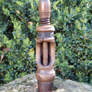 May include: An antique wooden corkscrew with a metal base and a large spring. The corkscrew is made of dark wood and has a cylindrical shape. The background is a green bush.