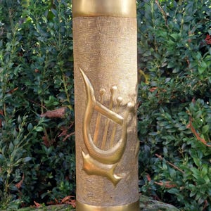 May include: A tall, cylindrical, gold-colored vase with a textured surface and a decorative lyre design. The vase has a crown-like top and a wide base. The background features green foliage.