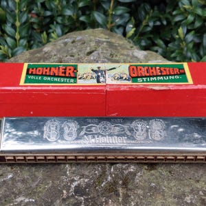 May include: A silver harmonica in its original red box. The box has the text "HOHNER" and "ORCHESTER" in black lettering. The harmonica has the text "M. HOHNER" in silver lettering.