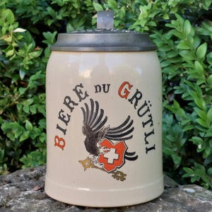 Könnte beinhalten: Cremefarbener Bierkrug aus Keramik mit Metalldeckel. Der Krug trägt die Worte "BIERE DU GRUTLI" in Rot und Schwarz sowie ein Adleremblem und ein Schild mit einem Kreuz.