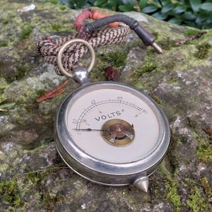 Puede incluir: Un voltímetro analógico vintage de color plateado con una esfera blanca y marcas negras. La esfera indica "VOLTS" y una aguja apunta al número 2. El voltímetro está conectado a un cordón trenzado marrón y negro con un conector rojo y negro.