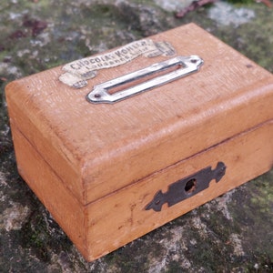 May include: A wooden box with a metal latch and a slot on the top. The box has a label that reads "Chocolats Kohler Lausanne Suisse".