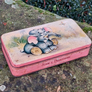 May include: A vintage pink tin box featuring a cartoon elephant illustration. The box is labelled "John S. Derbyshire & Son Limited, Nottingham, England".