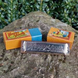 May include: A vintage harmonica in a brown box with the words "Happy Youth Harmonica" printed on it. The harmonica is silver with the words "Happy Youth" engraved on it. The box is sitting on a rock with green foliage in the background.