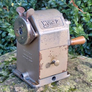 Peut inclure: Un taille-crayon Wyna 2 vintage avec une finition dorée métallique. Le taille-crayon a une poignée en bois et une manivelle. Le texte "Wyna 2" est visible en haut. Le taille-crayon est posé sur une roche.