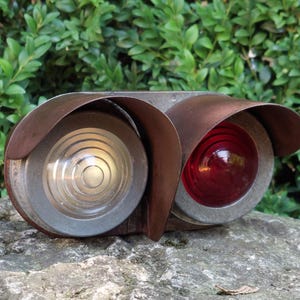 May include: An antique, dual-lens railroad signal light. One lens is clear, the other is red. The lenses are set in a weathered metal housing with a curved, rusty-brown visor above each lens. The light sits on a stone surface against a backdrop of green foliage.