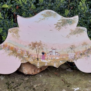 Può includere: Un decoro da parete in legno rosa con una scena dipinta di un piccolo villaggio con una donna e un bambino che camminano in lontananza. Il decoro ha la forma di uno stendardo con una parte superiore curva e due lati curvi.
