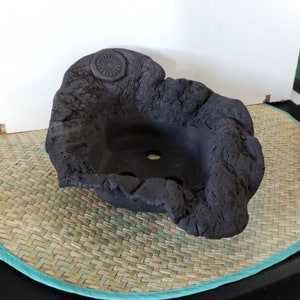 以下が含まれることがあります： 黒色のテクスチャ加工が施された粗い表面のセラミックプランター。プランターの上部に円形の開口部があり、下部に排水穴がいくつかあります。