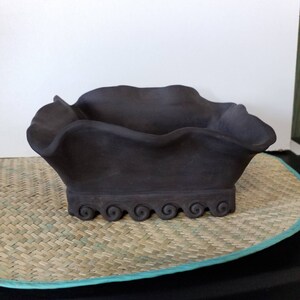 May include: A dark brown ceramic bowl with a wavy rim and a decorative base. The bowl is sitting on a woven mat.