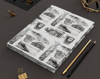 Puede incluir: Un diario blanco con un patrón de boceto en blanco y negro de arquitectura griega y búhos. Lápices dorados y negros, clips dorados y un cuadrado negro sobre una superficie gris oscuro.