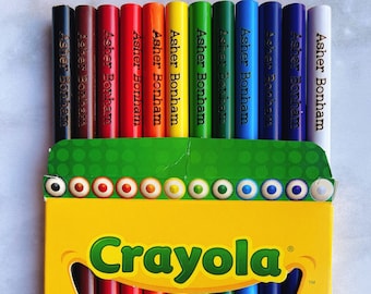 May include: A yellow Crayola box containing coloured pencils, each engraved with the name Asher Bonham. The pencils are arranged in a rainbow spectrum, featuring black, brown, red, orange, yellow, green, blue, purple, and white colours.