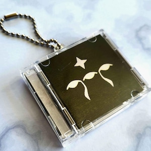 May include: A clear plastic CD case keychain with a silver ball chain. The case holds a black insert with a white star and two stylized white leaf shapes. The keychain is on a light blue and white marble surface.