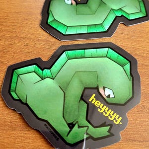 May include: Two green stickers featuring a stylized cartoon alligator with the text "heyyyy." The alligator has a geometric design with a black outline and a light green accent. The stickers are on a wooden surface.