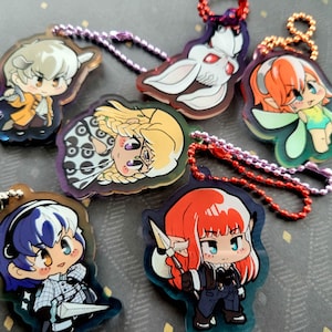 May include: A selection of colourful anime-style acrylic keychains. Each keychain features a unique character design with vibrant colours and attached metal ball chains. The characters have large eyes and are in various poses.