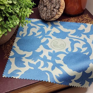 May include: A square, blue and white patterned fabric with a floral design. The fabric has a light blue background with darker blue floral shapes. A small, circular emblem is in the center. The fabric is placed on a dark red book.