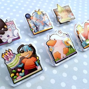 May include: A collection of seven clear acrylic pins featuring cartoon animal characters. The pins include a black cat, a blue elephant, a person in a green hat, a pink penguin, a duck, and a dog holding a cake. The background is a light blue surface with white polka dots.
