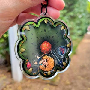 May include: A clear acrylic keychain featuring a cartoon character with blonde hair and a green, red, and black background. The character is holding a sword and shield.