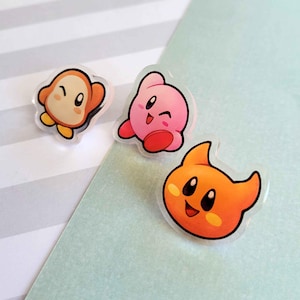 May include: Three colourful cartoon character pins. A pink Kirby character, an orange Waddle Dee character, and a yellow King Dedede character.