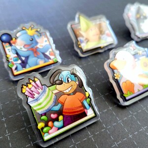 May include: A collection of cartoon character acrylic pins. One pin features a blue dog holding a birthday cake with candles. Other pins depict a blue koala, a duck, and a character in a green hat. The pins have a clear, glossy finish.