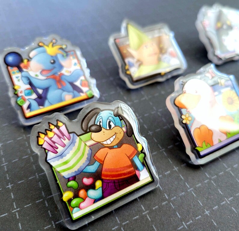 May include: A collection of cartoon character acrylic pins. One pin features a blue dog holding a birthday cake with candles. Other pins depict a blue koala, a duck, and a character in a green hat. The pins have a clear, glossy finish.