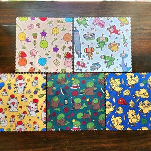 Peut inclure: Six carrés de tissu colorés avec des motifs répétitifs. Les motifs incluent des robots de dessins animés, des chats et divers fruits et légumes. Les tissus sont dans les tons de jaune, bleu, vert et crème, sur un fond en bois foncé.