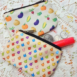 May include: Two small zippered pouches with different patterns. One pouch has a beige background with a repeating pattern of colourful cartoon characters. The other pouch has a beige background with a repeating pattern of vegetables and pumpkins.