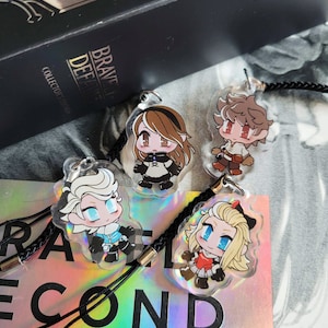 May include: Four colourful anime character charms with black braided cords. The charms are made of clear plastic with a holographic finish. The characters are a girl with white hair, a girl with brown hair, a girl with blonde hair, and a boy with brown hair.