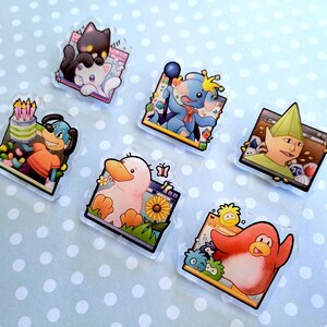 May include: A collection of colorful, cartoon-style acrylic pins featuring various characters. Designs include a black cat on a white cat, a blue dog with a birthday cake, a blue elephant-like creature, a duck with a sunflower, a green-hatted character, and a red penguin.