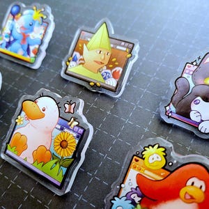 May include: A collection of clear acrylic pins featuring cartoon characters. Designs include a blue elephant, a duck with a sunflower, a penguin, a dog, a cat, and a character with a green hat. Each pin has a unique design and color scheme.