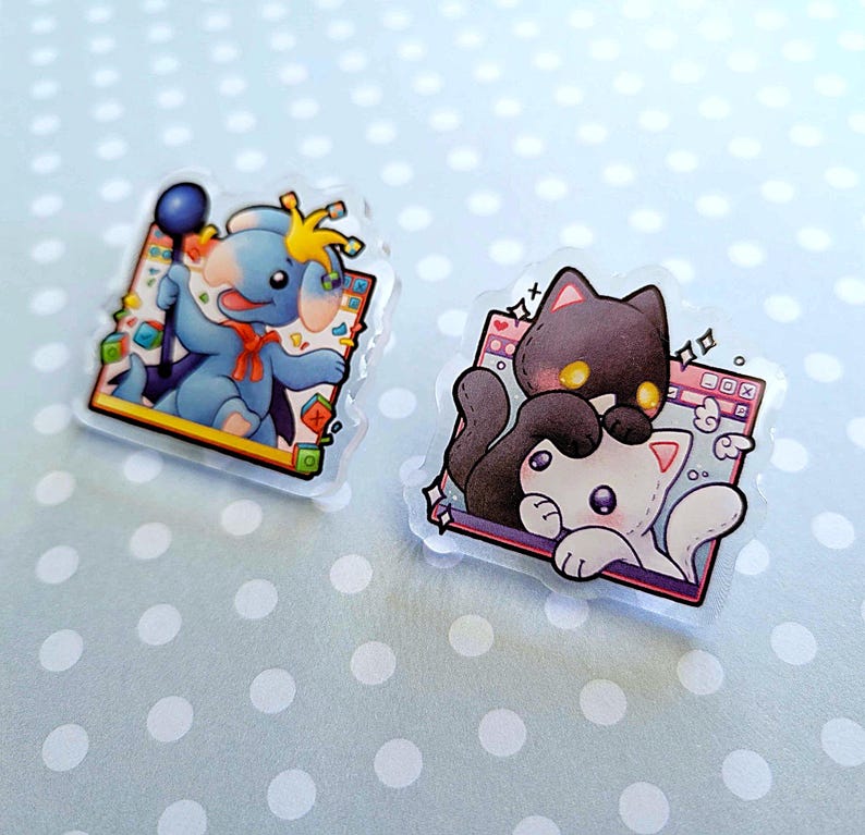 May include: Two clear acrylic pins featuring cartoon characters. One pin shows a blue elephant with a crown and scepter. The other pin depicts a black cat and a white cat. Both pins have a colorful border.