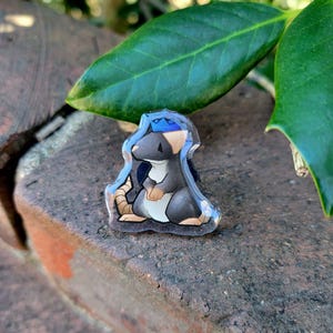 May include: A clear acrylic pin featuring a cartoon rat wearing a blue crown. The rat is gray, black, and white with tan accents. The pin is set against a textured brick surface with green leaves in the background.