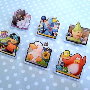 May include: A collection of six clear acrylic pins featuring cartoon characters. The pins showcase a variety of designs, including a black cat and white cat, a blue creature with a staff, a green elf, a dog with a cake, a duck with a sunflower, and a red penguin.