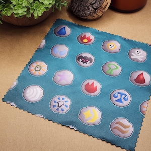 May include: A teal square cloth featuring a repeating pattern of stylized, colorful icons resembling runes or symbols on gray stones. The icons include water drops, flames, and other fantasy-themed designs. The cloth is placed on a brown surface.