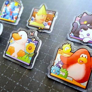 May include: A collection of clear acrylic pins featuring cartoon characters. The pins showcase a variety of designs, including a duck with a sunflower, a penguin, and a cat. The pins have a glossy finish and are set against a dark background.
