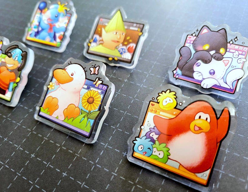 May include: A collection of clear acrylic pins featuring cartoon characters. The pins showcase a variety of designs, including a duck with a sunflower, a penguin, and a cat. The pins have a glossy finish and are set against a dark background.