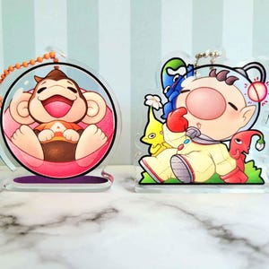 May include: Two acrylic keychains featuring cartoon characters. One shows a smiling monkey inside a clear bubble, the other depicts a character in a yellow spacesuit with colorful companions. Both have a metal chain for attachment.