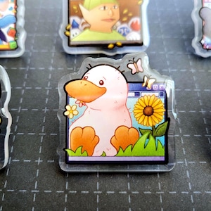May include: A clear acrylic charm featuring a cartoon duck with a pink body, orange beak and feet, and a sunflower. The duck is set against a computer screen background with butterflies and flowers.