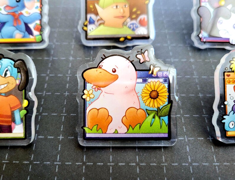 May include: A clear acrylic charm featuring a cartoon duck with a pink body, orange beak and feet, and a sunflower. The duck is set against a computer screen background with butterflies and flowers.