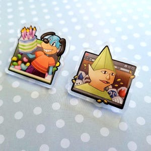 May include: Two clear acrylic pins. One features a cartoon dog holding a cake with candles. The other pin shows a cartoon character with a green hat and a computer screen background. Both pins have a black border.