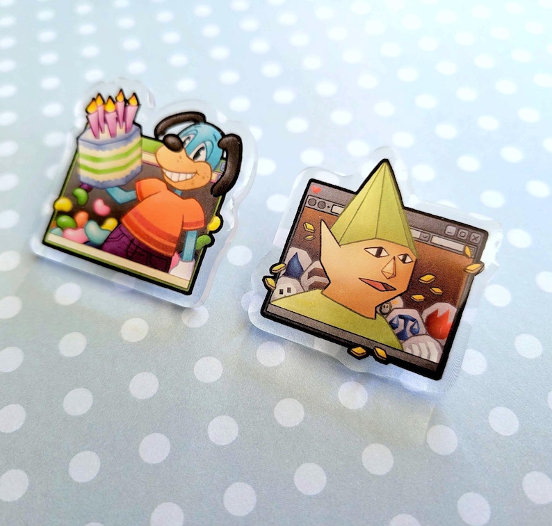 May include: Two clear acrylic pins. One features a cartoon dog holding a cake with candles. The other pin shows a cartoon character with a green hat and a computer screen background. Both pins have a black border.